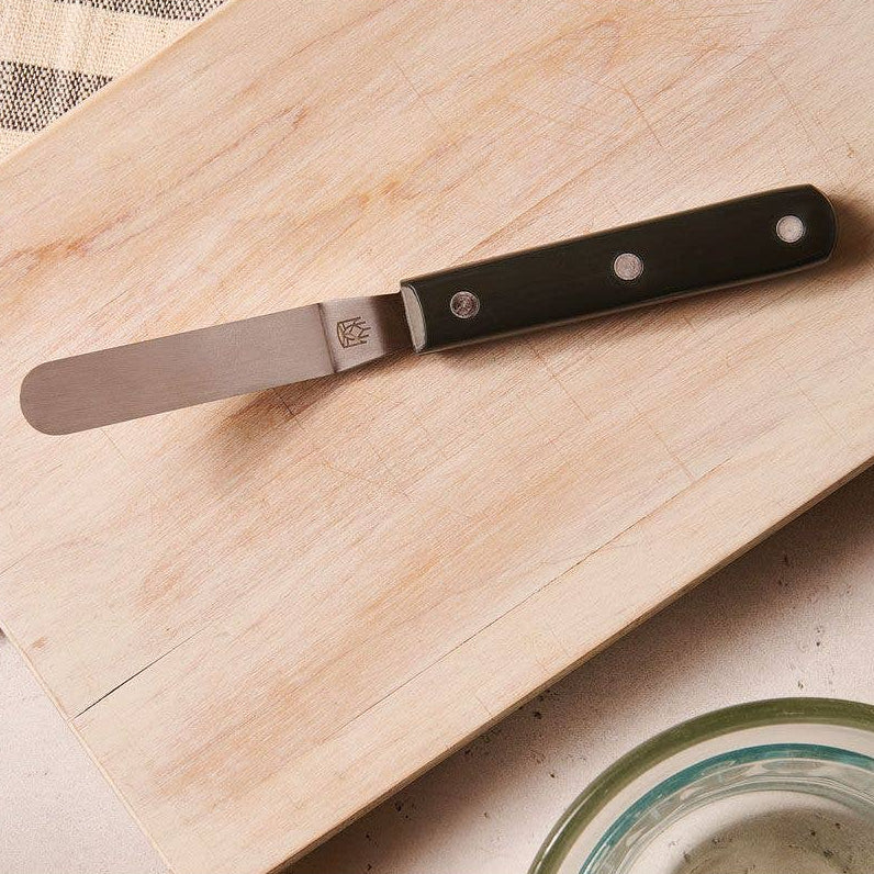 Spreader knife on a wooden cutting board with a kitchen towel and glasses in the background