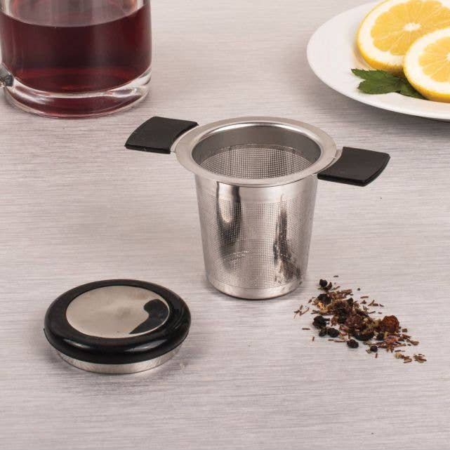 Stainless steel tea infuser on a table with tea leaves and a glass of tea.