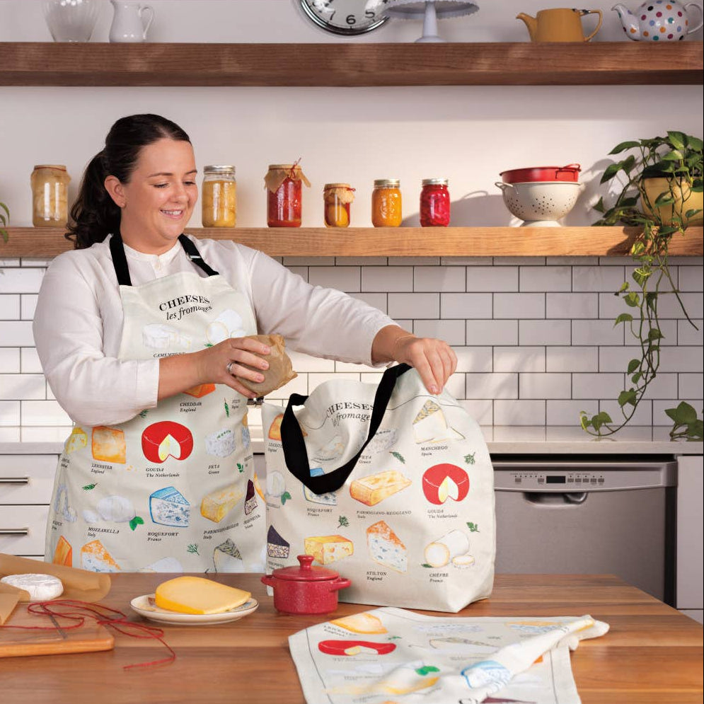 Woman in a kitchen holding a tote bag with food illustrations, wearing a matching apron.