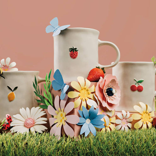 Three ceramic mugs with floral and fruit designs on a grassy surface with flowers and a lemon.