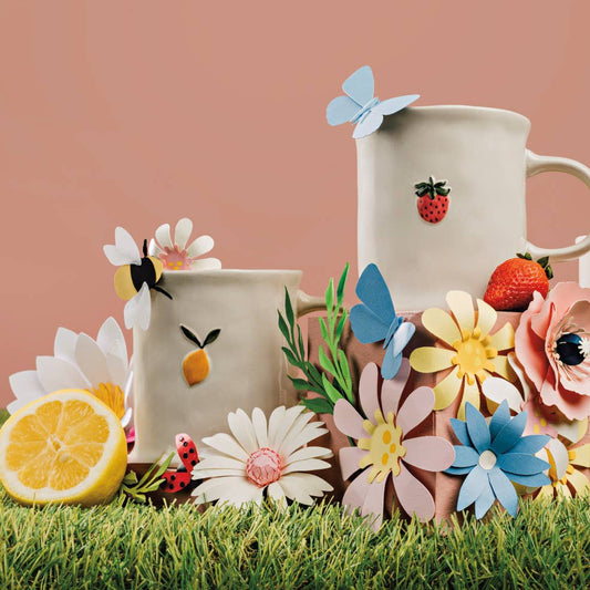 Three ceramic mugs with floral and fruit designs on a grassy surface with flowers and a lemon.