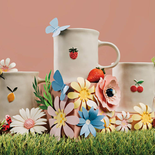 Three ceramic mugs with floral and fruit designs on a grassy surface with flowers and a lemon.