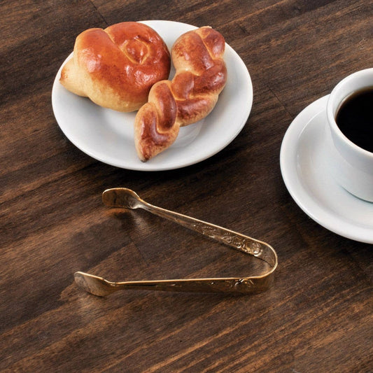 Two pastries on a white plate with a cup of coffee and gold tongs on a wooden surface.