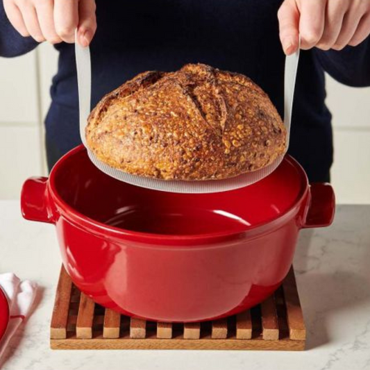 Hands using a white loaf lifter to lift a loaf of bread out of a red dutch oven
