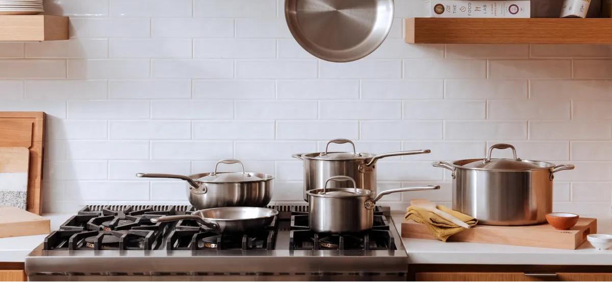 Stainless steel pots and pans on a gas stove with a tiled wall and wooden shelves in the background.