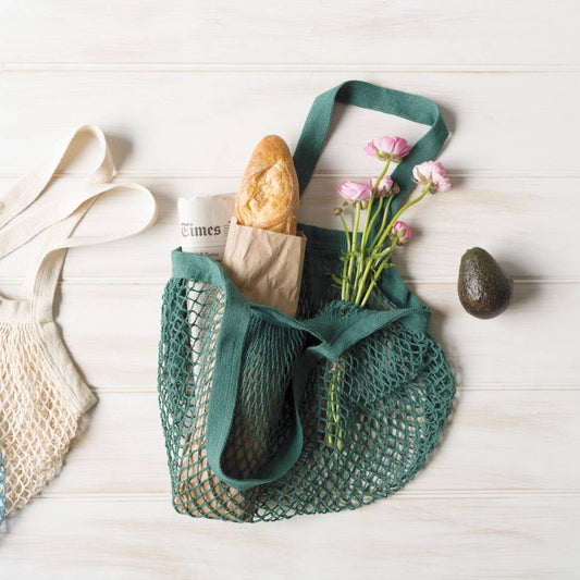 Green mesh bag with bread, flowers, and avocado on a wooden surface
