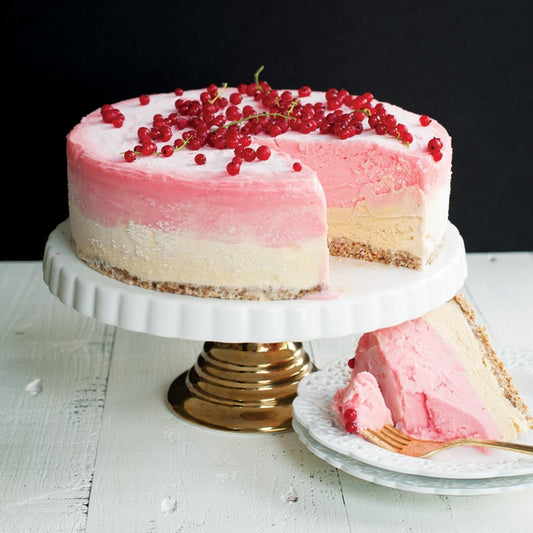 Pink and white cake with red berries on a white stand, slice taken out, on a white surface.