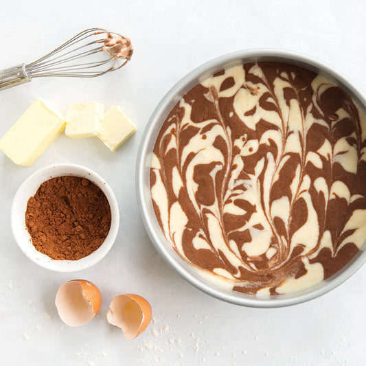 Baking ingredients including a marbled cake batter in a pan, cocoa powder, butter, and eggs on a white surface.
