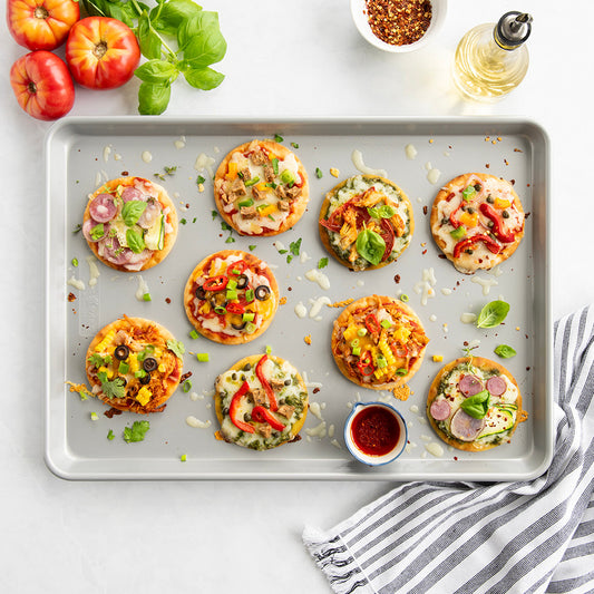 Sheet pan with mini pizzas on a white surface with tomatoes and basil.