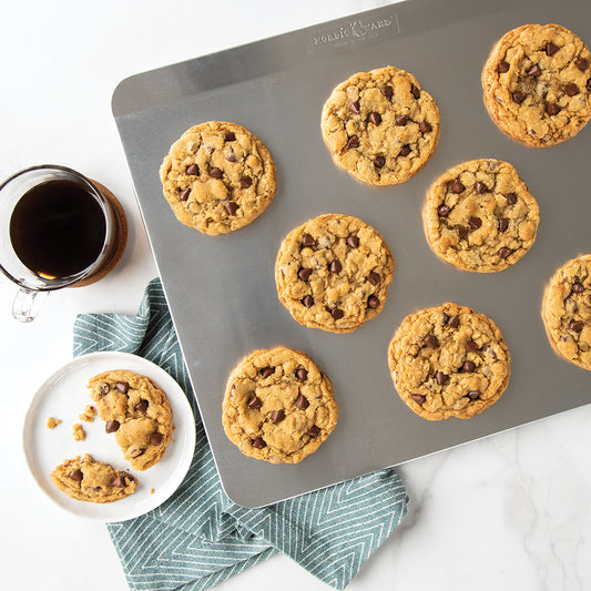 Chocolate chip cookies on a baking tray with a cup of coffee on a white surface.