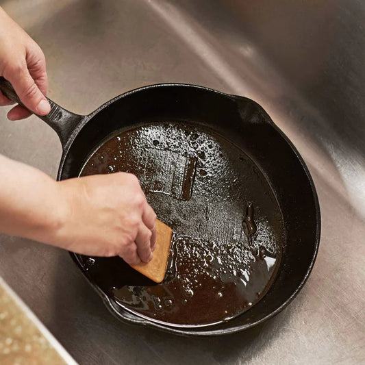 Person cleaning a cast iron skillet with a wooden scraper in a kitchen sink.