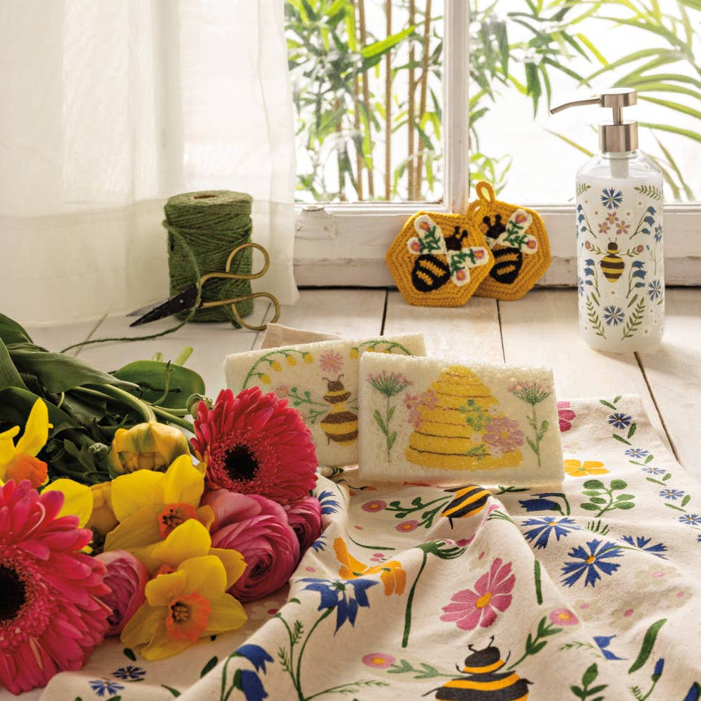 Decorative tablecloth with floral and bee patterns on a table with flowers and a window in the background.