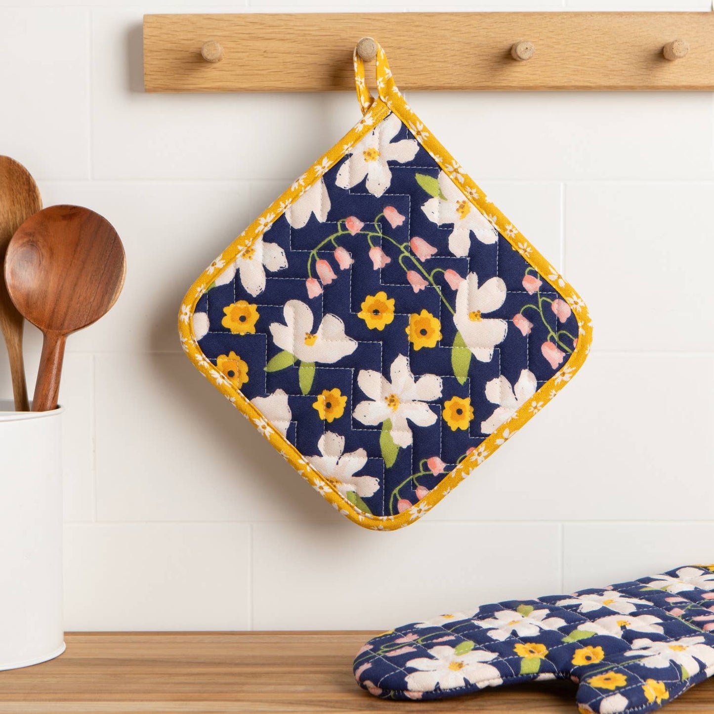 Floral patterned pot holder and oven mitt hanging on a wooden rack with a white tiled wall background.
