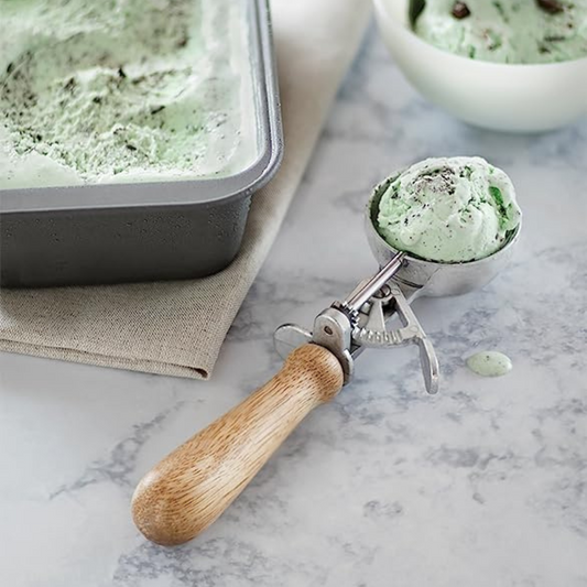 Green ice cream in a metal scoop with wooden handle on a marble surface.