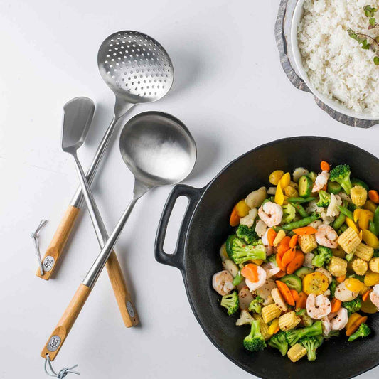 Wok skimmer tools stainless steel with wooden handles on white surface next to a black wok with fresh vegetables and bowl of white rice