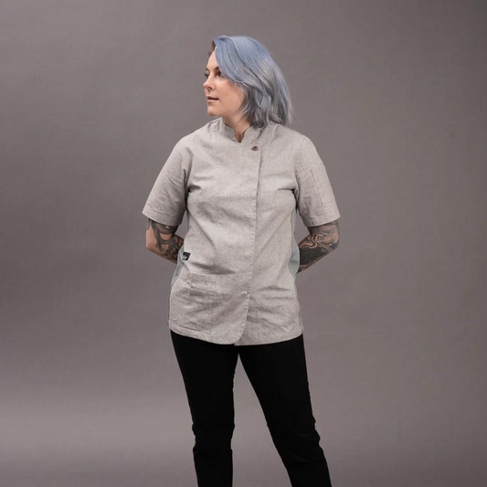A woman chef wearing a medium-sized, gray, short-sleeved chef coat with a round neckline, buttoned front, and a pocket on the left side. She is also wearing black pants and has light blue hair.
