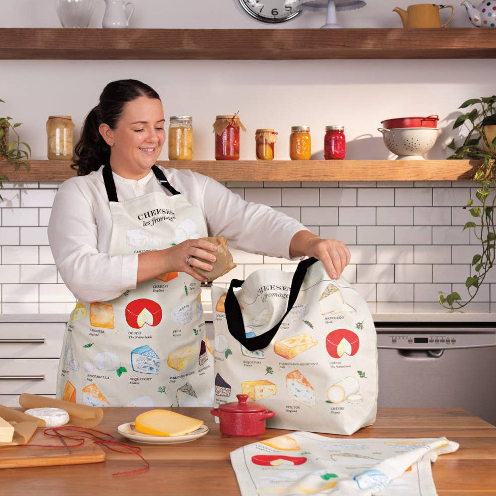 Woman in a kitchen holding a tote bag with food illustrations, wearing a matching apron.