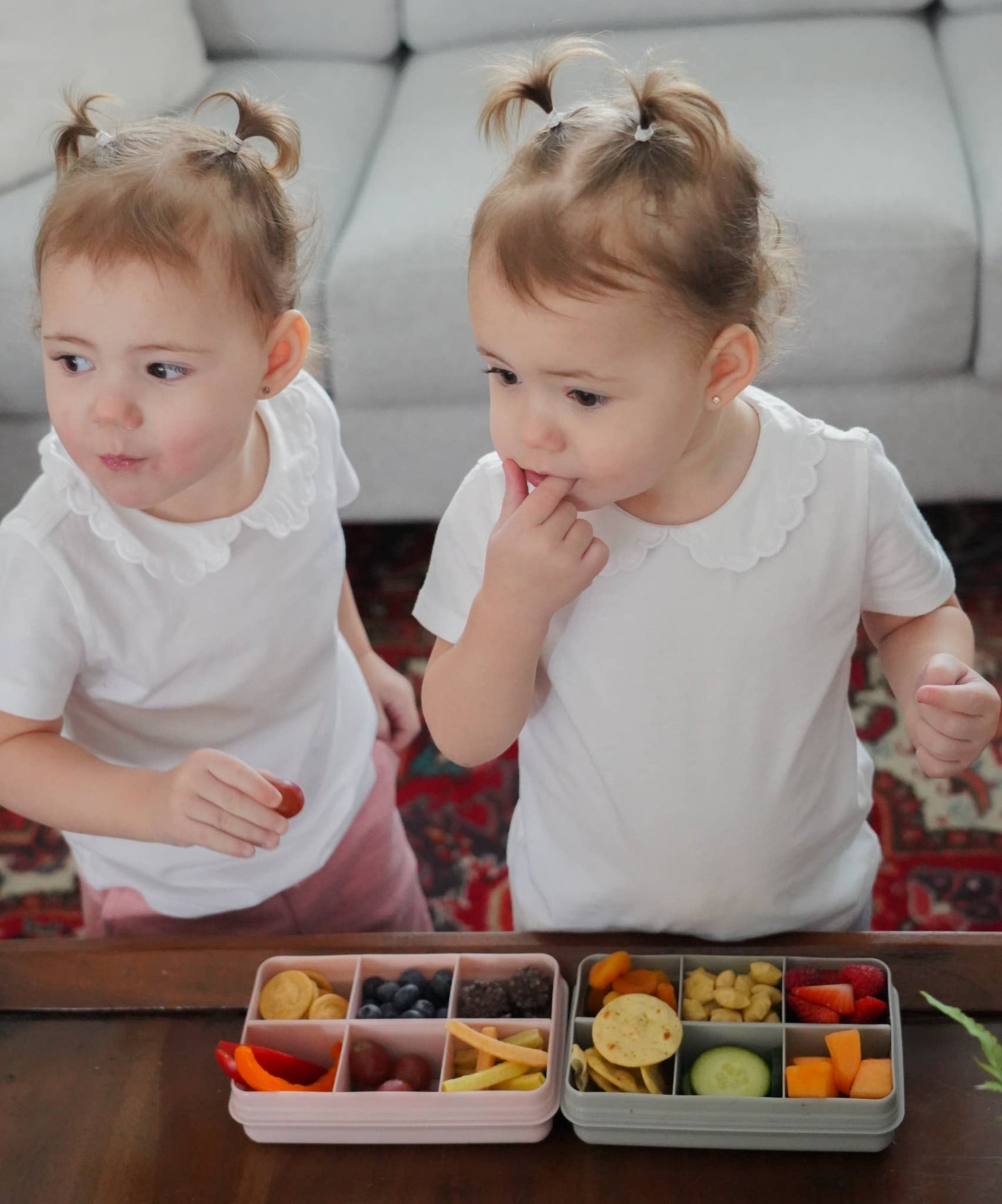Two young girls sitting at a table with divided snack containers filled with various food items.