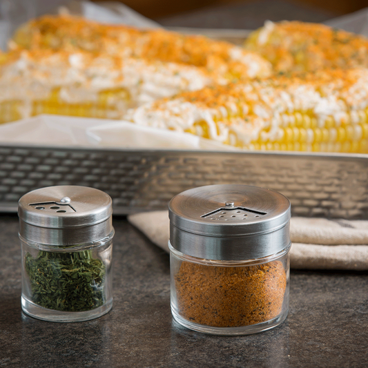 Two spice containers with a dish of corn on the cob in the background.