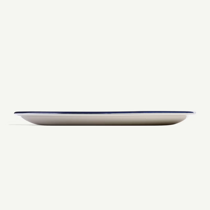 A long, shallow oval plate in warm off-white with a thin dark blue rim, shown from a side profile against a solid off-white background.