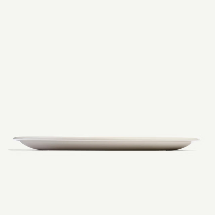 A minimalist, shallow oval plate in a matte light beige or off-white finish, shown from a side profile against a solid off-white background.