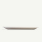 A minimalist, shallow oval plate in a matte light beige or off-white finish, shown from a side profile against a solid off-white background.