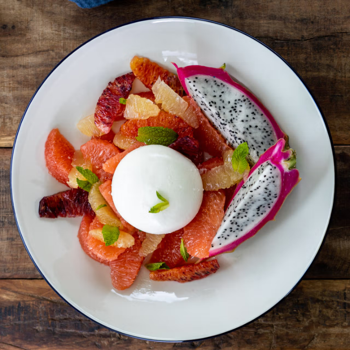 A top-down view of a white plate with a dark rim, filled with a colorful fruit salad featuring grapefruit, dragon fruit, mandarin, and a white berry or gelato center, set on a rustic wooden table.