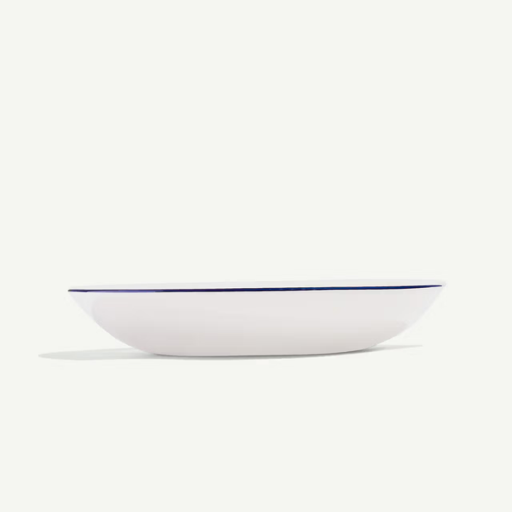 A white oval serving bowl with a thin dark blue rim, photographed from a side angle against a clean off-white background.