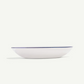 A white oval serving bowl with a thin dark blue rim, photographed from a side angle against a clean off-white background.
