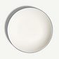 A top-down view of a round white ceramic plate with a thin dark blue rim, set against a clean off-white background with a soft shadow.