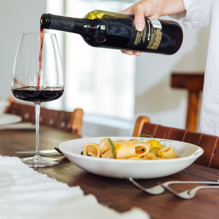 A person pouring red wine from a dark bottle into a clear wine glass on a wooden dining table, with a plate of pasta in the foreground.