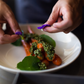 A close-up of a plated dish of cooked baby carrots topped with green herb sauce, served in a white bowl with a person's hands placing purple edible flowers on top.
