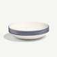 A stack of five white ceramic bowls with a thin dark blue rim, photographed on a clean off-white background with a soft shadow.