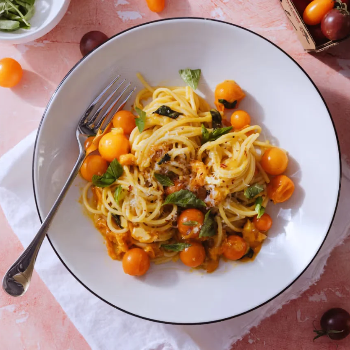 A top-down view of a white bowl with a thin black rim, filled with spaghetti in a creamy orange sauce and topped with halved cherry tomatoes and fresh basil leaves. A silver fork rests on the left side of the bowl, set on a white napkin over a pink surface.