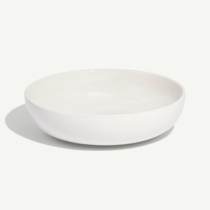 A minimalist white ceramic bowl with a shallow, wide rim, shown from a side angle on a clean white background with a soft shadow.