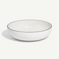 A white ceramic bowl with a thin black rim, photographed on a clean off-white background with a soft shadow.