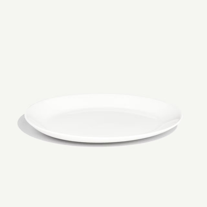 A minimalist white ceramic plate with a shallow rim, photographed from a high angle on a light off-white background with a soft shadow.