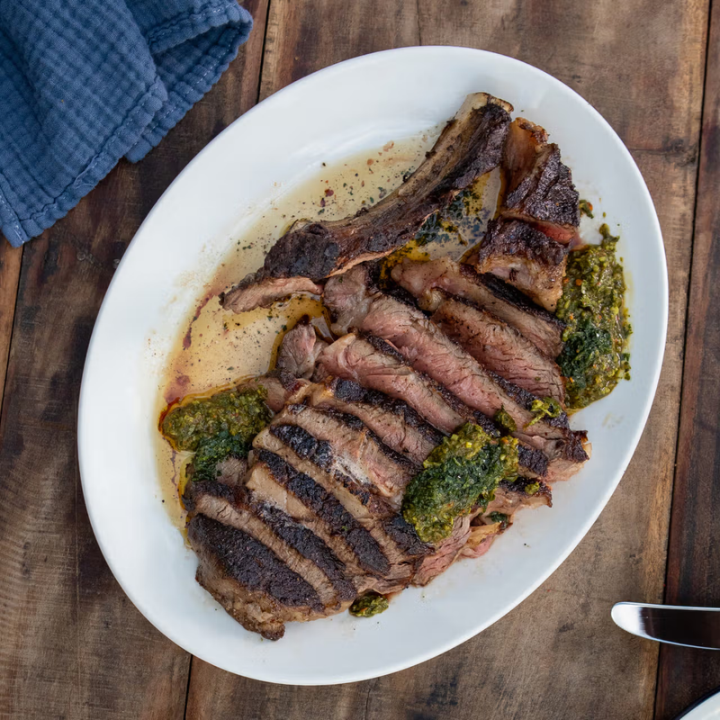 A plated sliced steak with green herb sauce on a white oval plate, set on a rustic wooden table with a blue cloth napkin and a knife partially visible.