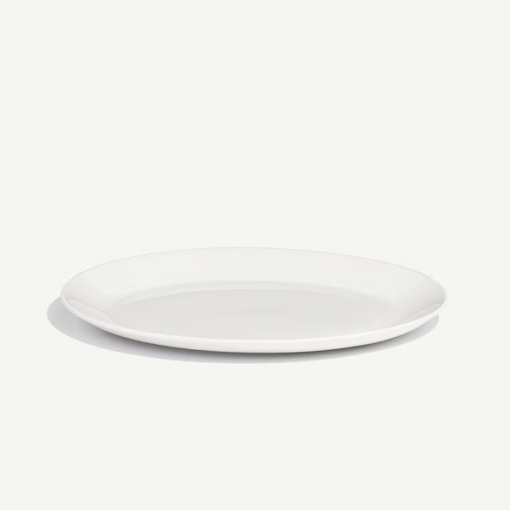 A minimalist white ceramic plate with a shallow rim, photographed from a high angle on a clean, off-white background with a soft shadow.