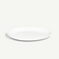A minimalist white ceramic plate with a shallow rim, photographed from a high angle on a light off-white background with a soft shadow.