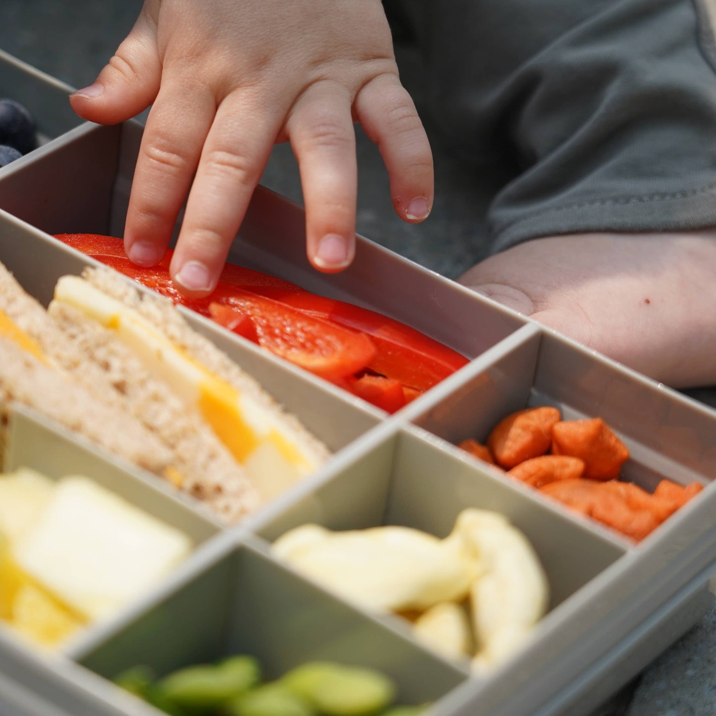 Child's hand reaching into a compartmentalized lunch box with various food items.