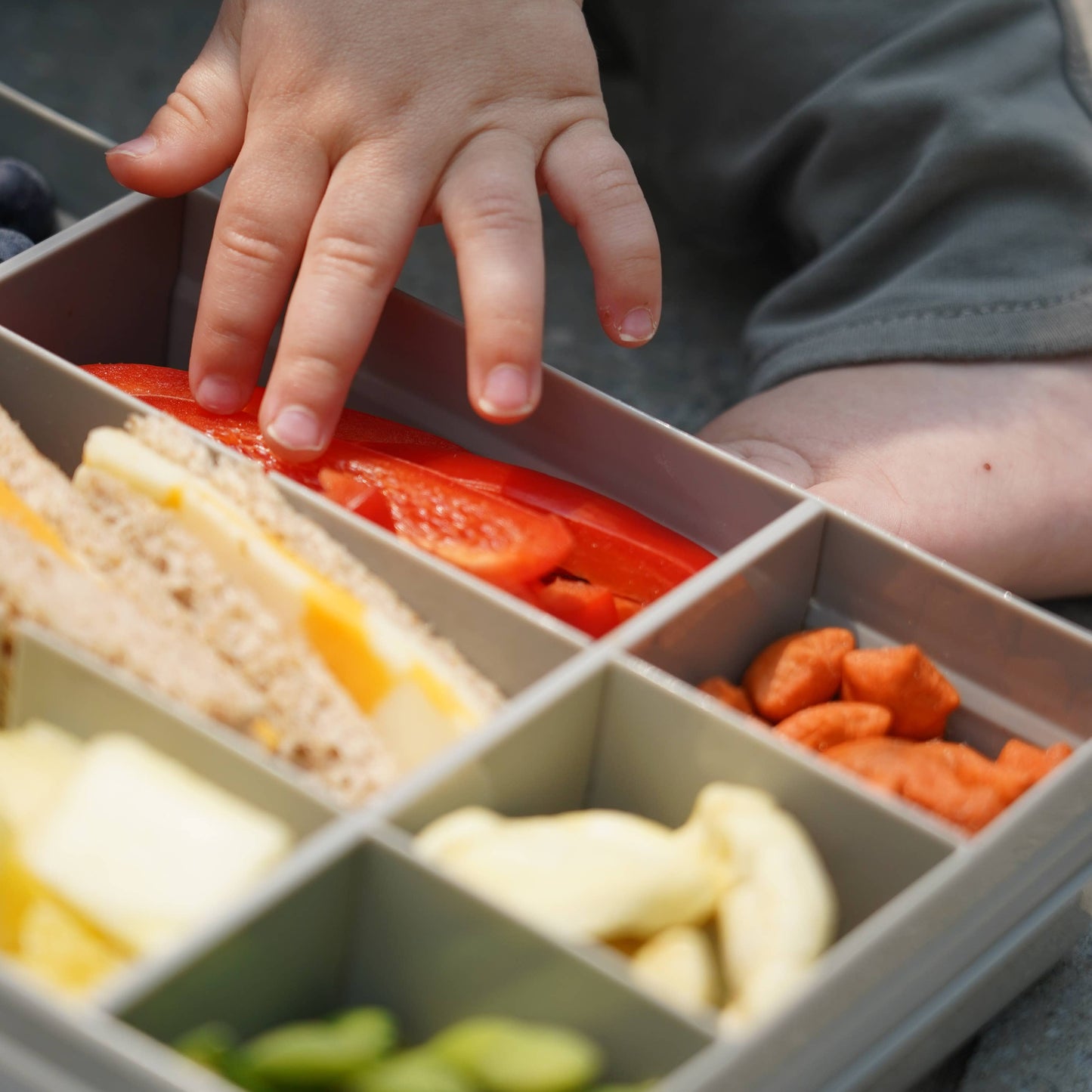 Child's hand reaching into a compartmentalized lunch box with various food items.