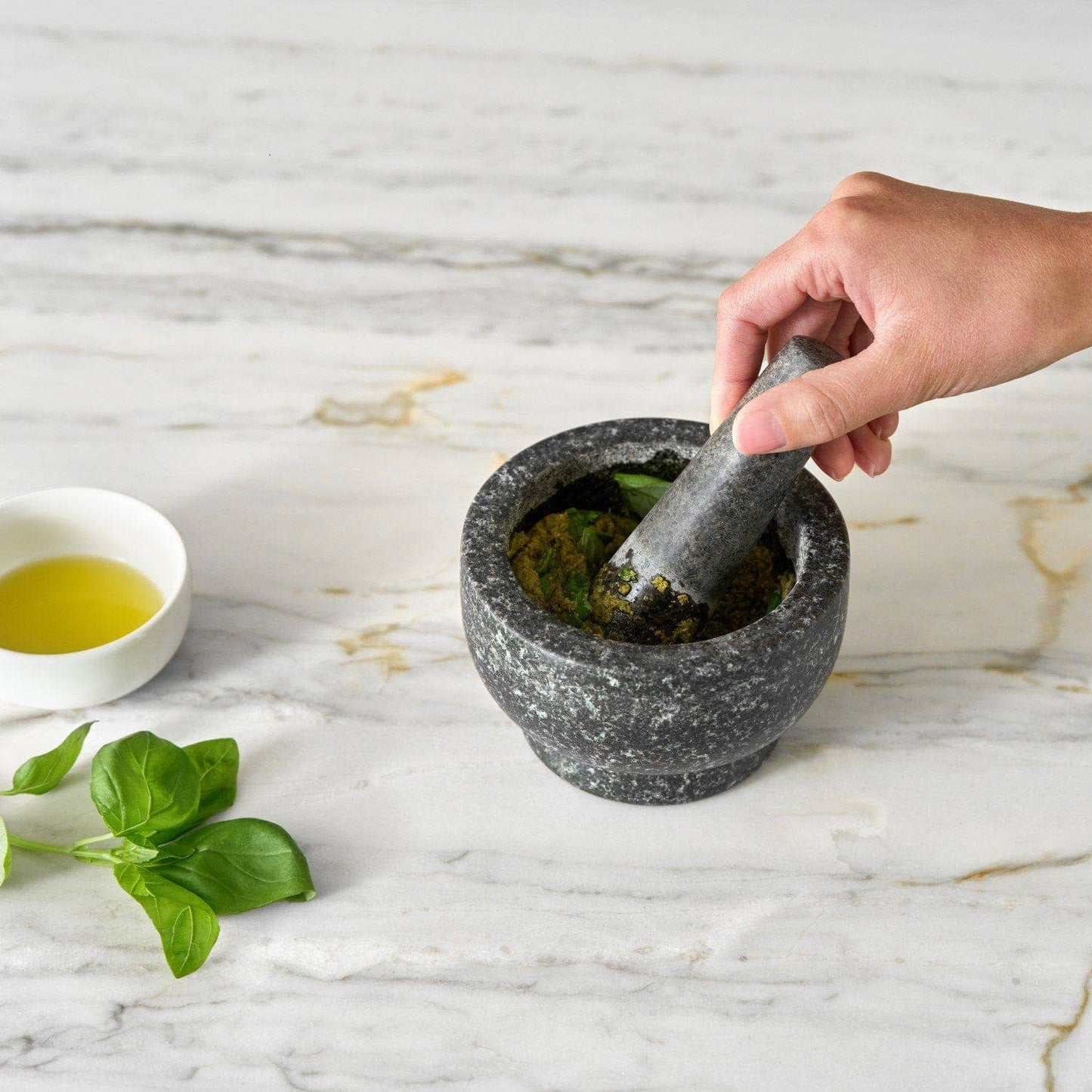 Person using a mortar and pestle on a marble surface with a small bowl of oil and leaves nearby.