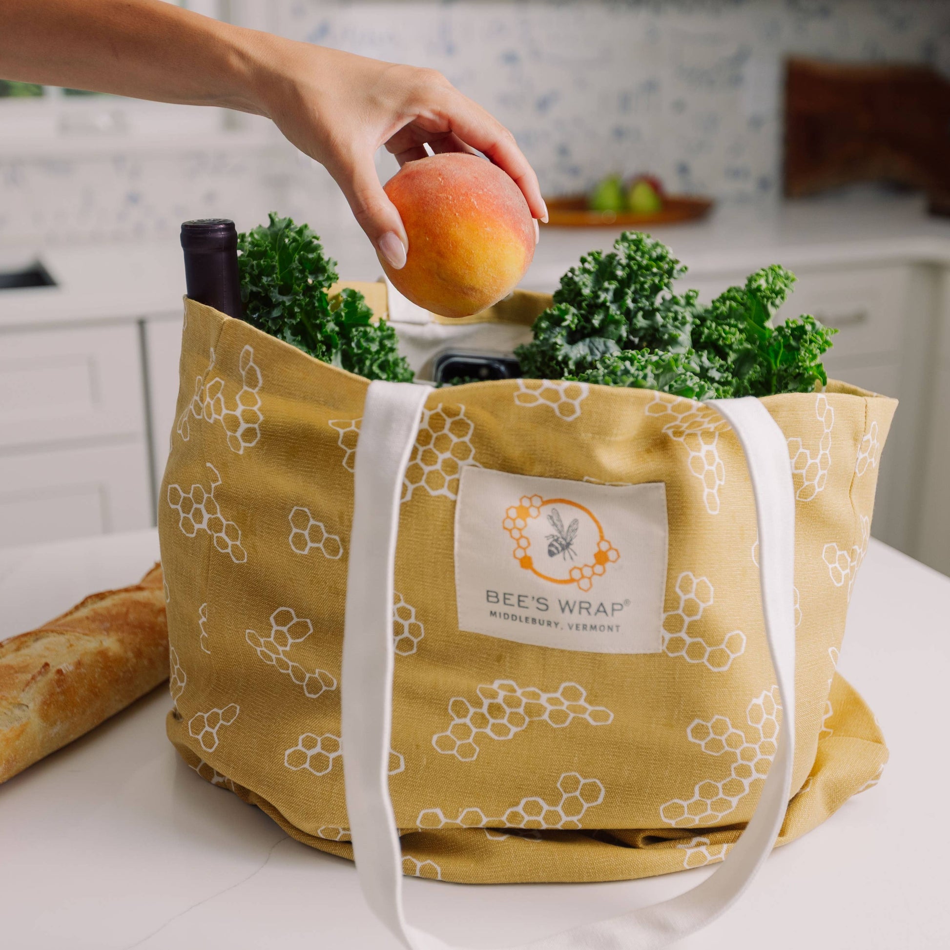 Yellow reusable shopping bag with produce and a hand holding an apple in a kitchen setting.