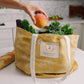 Yellow reusable shopping bag with produce and a hand holding an apple in a kitchen setting.