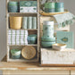 Display of green kitchenware including bowls, cups, and a towel on a wooden shelf.