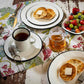 Breakfast setting with crumpets, tea, coffee, and fruit on a wooden table.