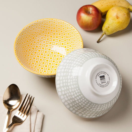 Two ceramic bowls with different patterns, a pear, apple, and cutlery on a light surface.