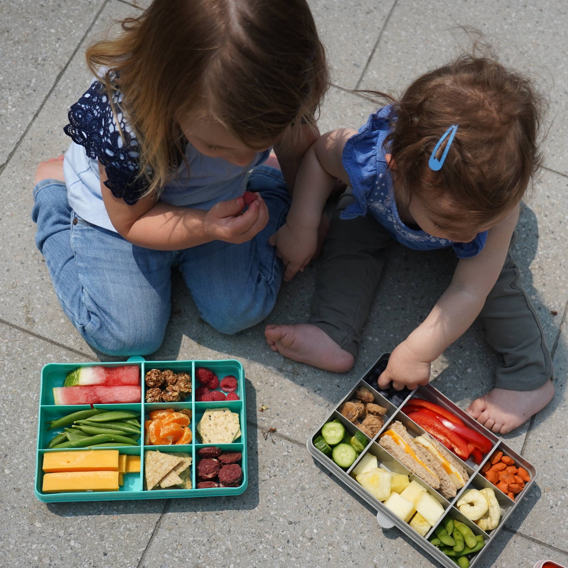 Two children sitting on a sidewalk with bento boxes filled with various food items.