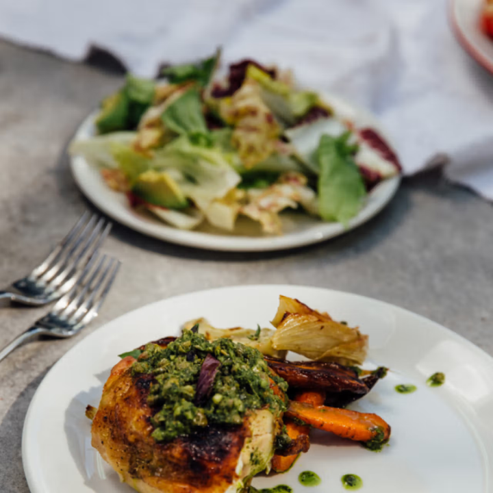 Plated dish with roasted chicken and vegetables on a table with a salad in the background.